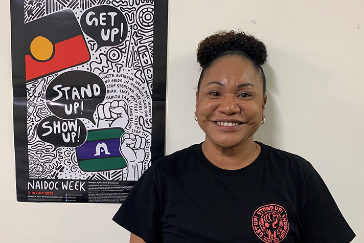 Hannah Ramsey, an Indigenous Australian lady smiles beautifully at the camera next to a poster for NAIDOC Week 2022
