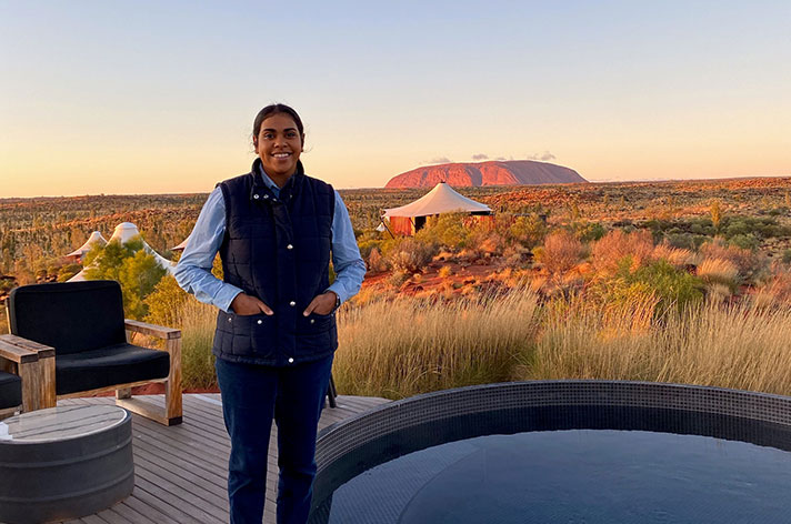 Young Indigenous woman Codie stands in an outdoor patio with a large circle bath, she smiles at the camera and Uluru sits on the horizon behind her. A glamping tent also sits in the background between Codie and Uluru. 